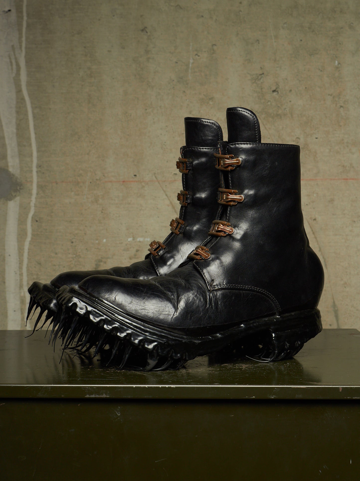 A pair of black leather combat boots with spiked soles and multiple brown buckle details, displayed on a dark surface against a textured concrete wall.
