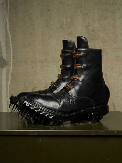 A pair of black leather combat boots with spiked soles and multiple brown buckle details, displayed on a dark surface against a textured concrete wall.