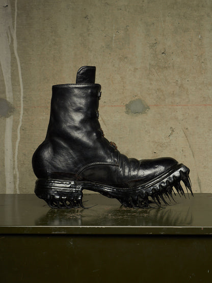 A black leather combat boot with a spiked, aggressive sole, displayed on a dark reflective surface against a textured concrete wall.