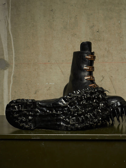 A black leather combat boot with three bronze-toned metal eyelets, resting on a dark surface against a textured concrete wall. The boot is heavily decorated with a glossy, highly textured black organic, claw-like or shell-like embellishment covering the toe and heel area.