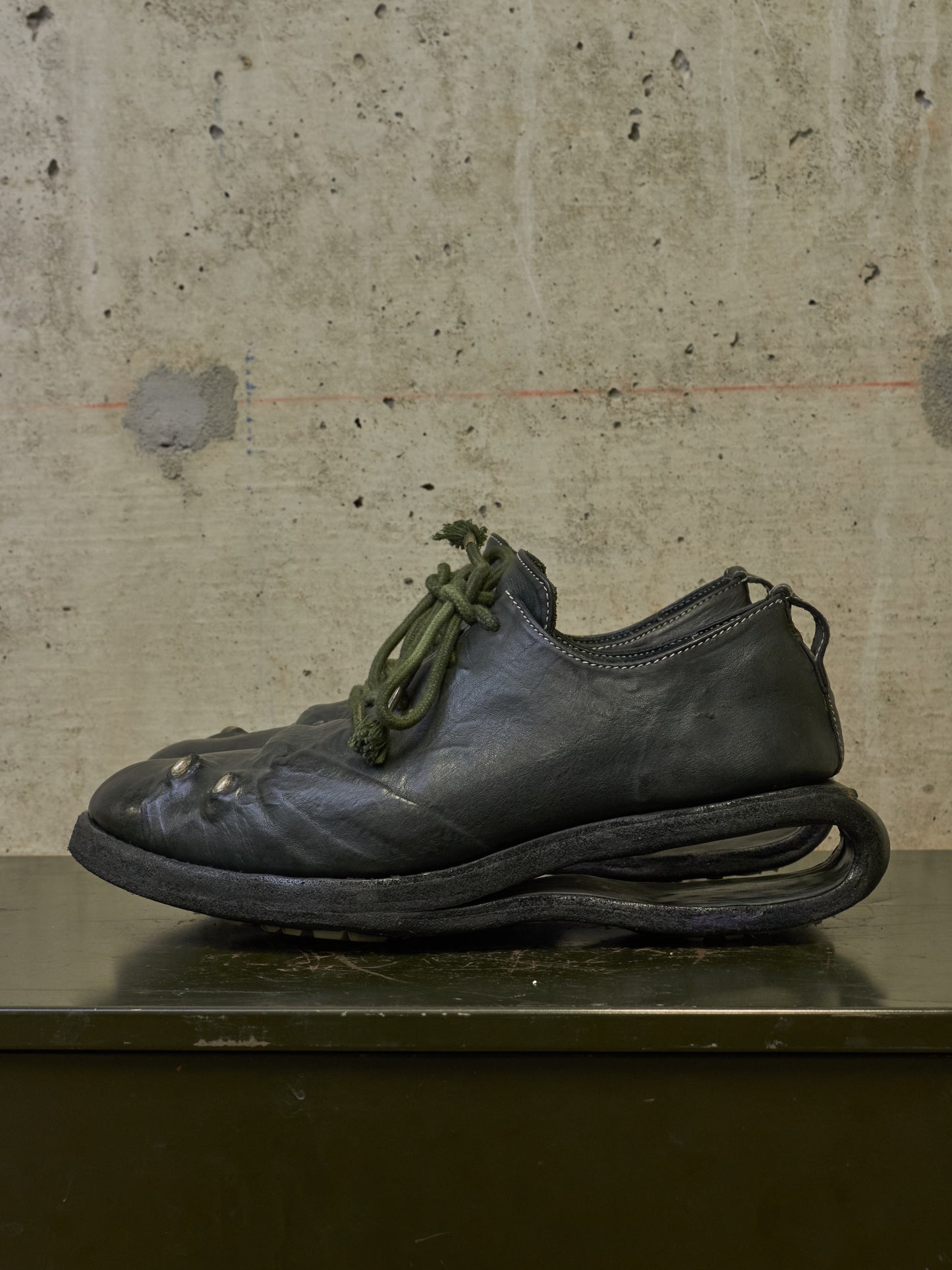 A single dark leather shoe with an unusual sculptural sole and olive green laces, photographed against a textured concrete wall on a dark surface.