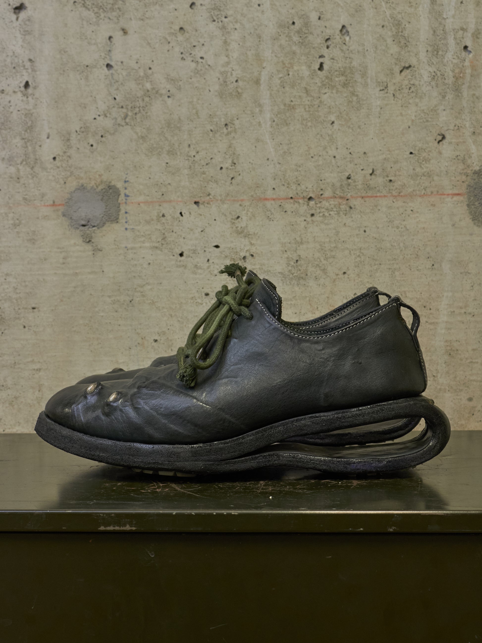 A single dark leather shoe with an unusual sculptural sole and olive green laces, photographed against a textured concrete wall on a dark surface.
