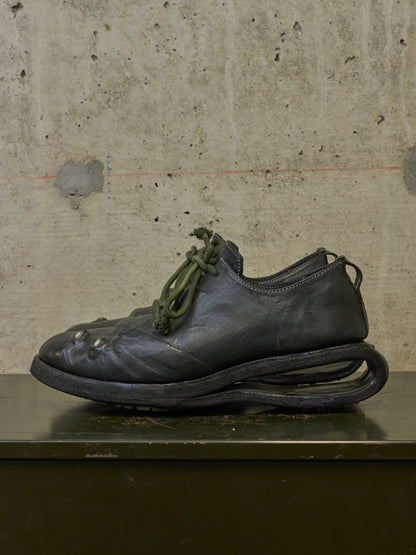 A single dark leather shoe with an unusual sculptural sole and olive green laces, photographed against a textured concrete wall on a dark surface.