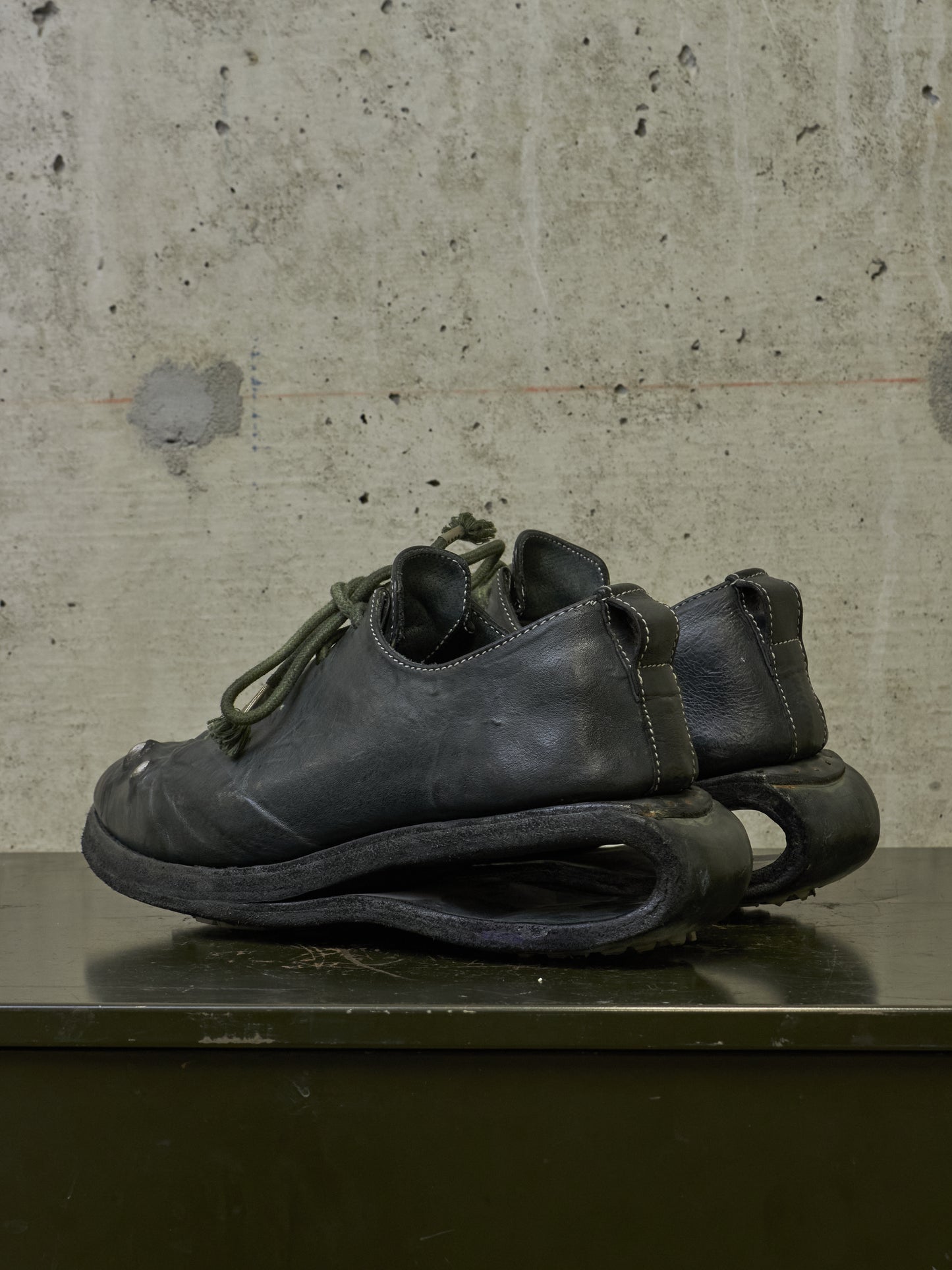 A pair of black leather low-top shoes with sculptural, curved soles resting on a dark surface against a textured concrete wall.