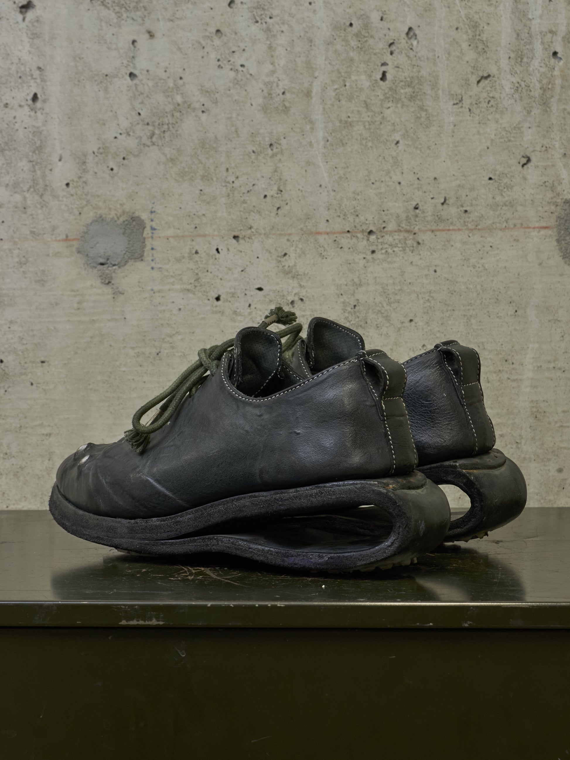 A pair of black leather low-top shoes with sculptural, curved soles resting on a dark surface against a textured concrete wall.