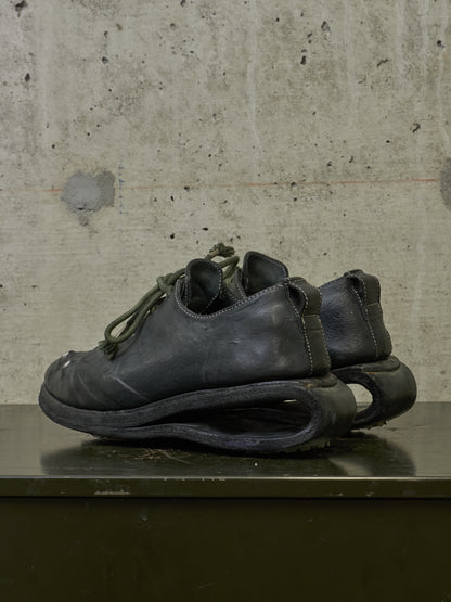 A pair of black leather low-top shoes with sculptural, curved soles resting on a dark surface against a textured concrete wall.