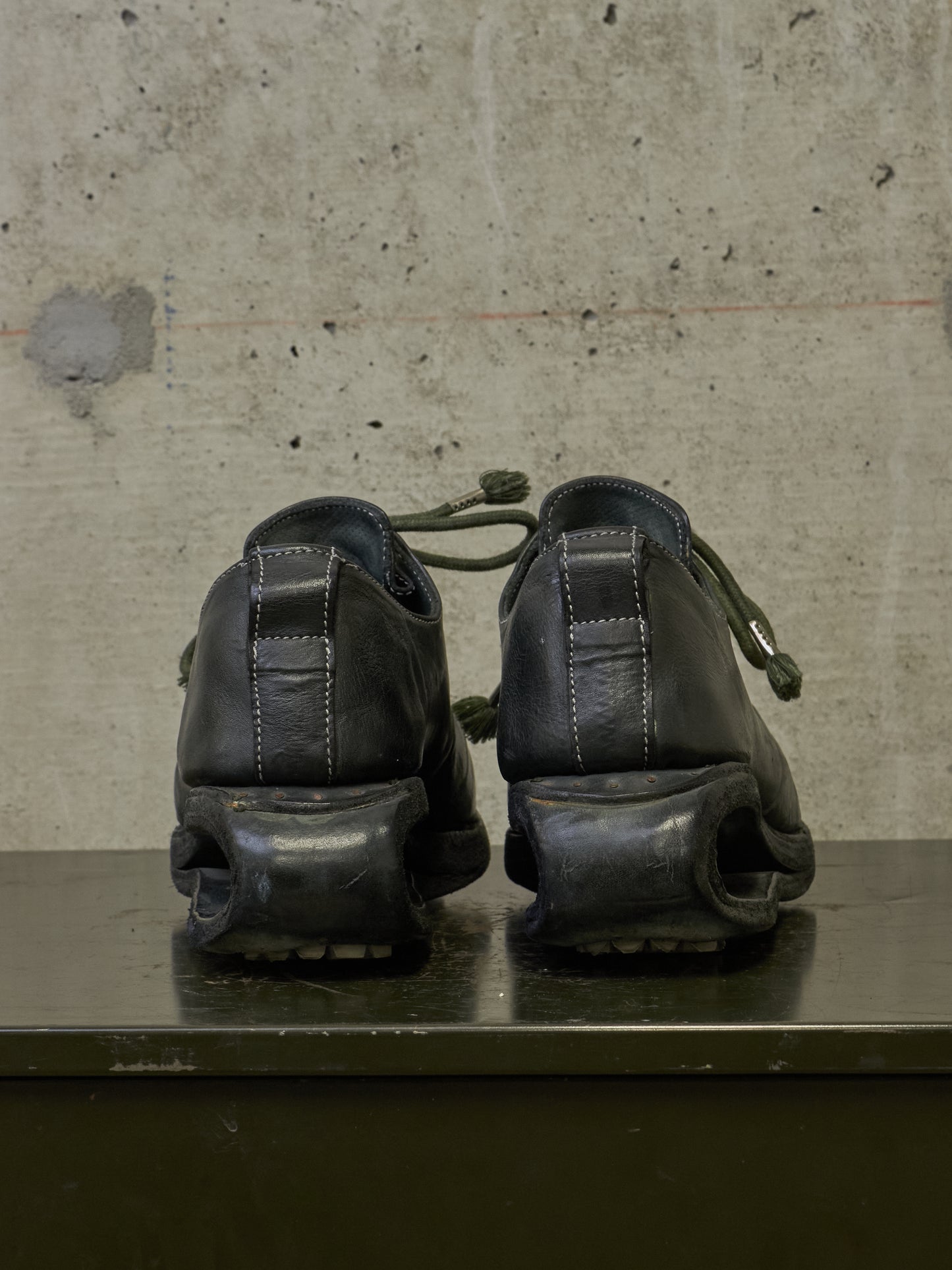 A pair of black leather platform shoes with chunky sculptural soles, shown from the back on a dark surface against a textured concrete wall.