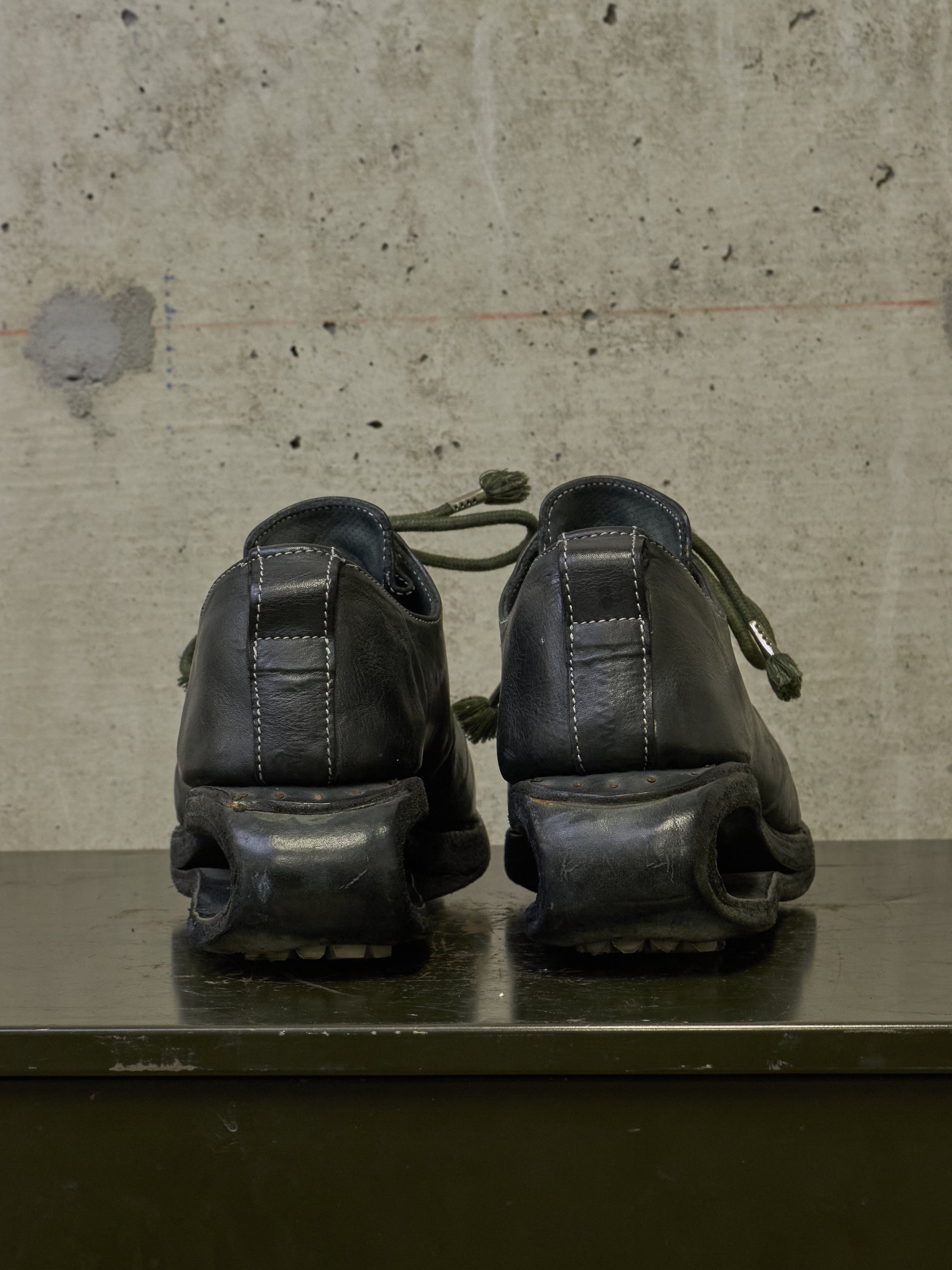 A pair of black leather platform shoes with chunky sculptural soles, shown from the back on a dark surface against a textured concrete wall.