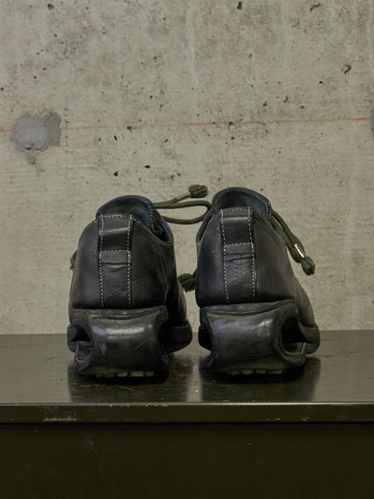 A pair of black leather platform shoes with chunky sculptural soles, shown from the back on a dark surface against a textured concrete wall.