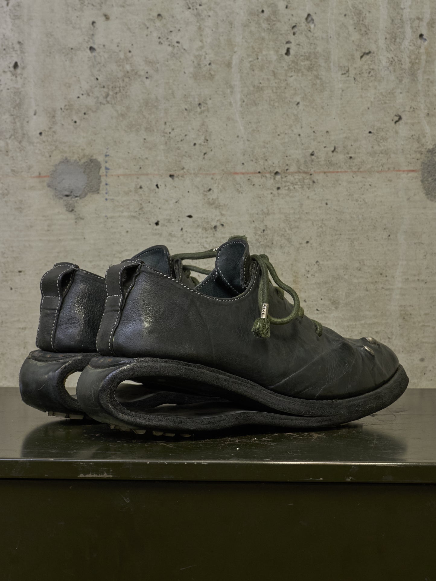 A pair of black leather shoes with a distinctive sculpted sole and olive green laces, displayed on a dark surface against a textured concrete wall.