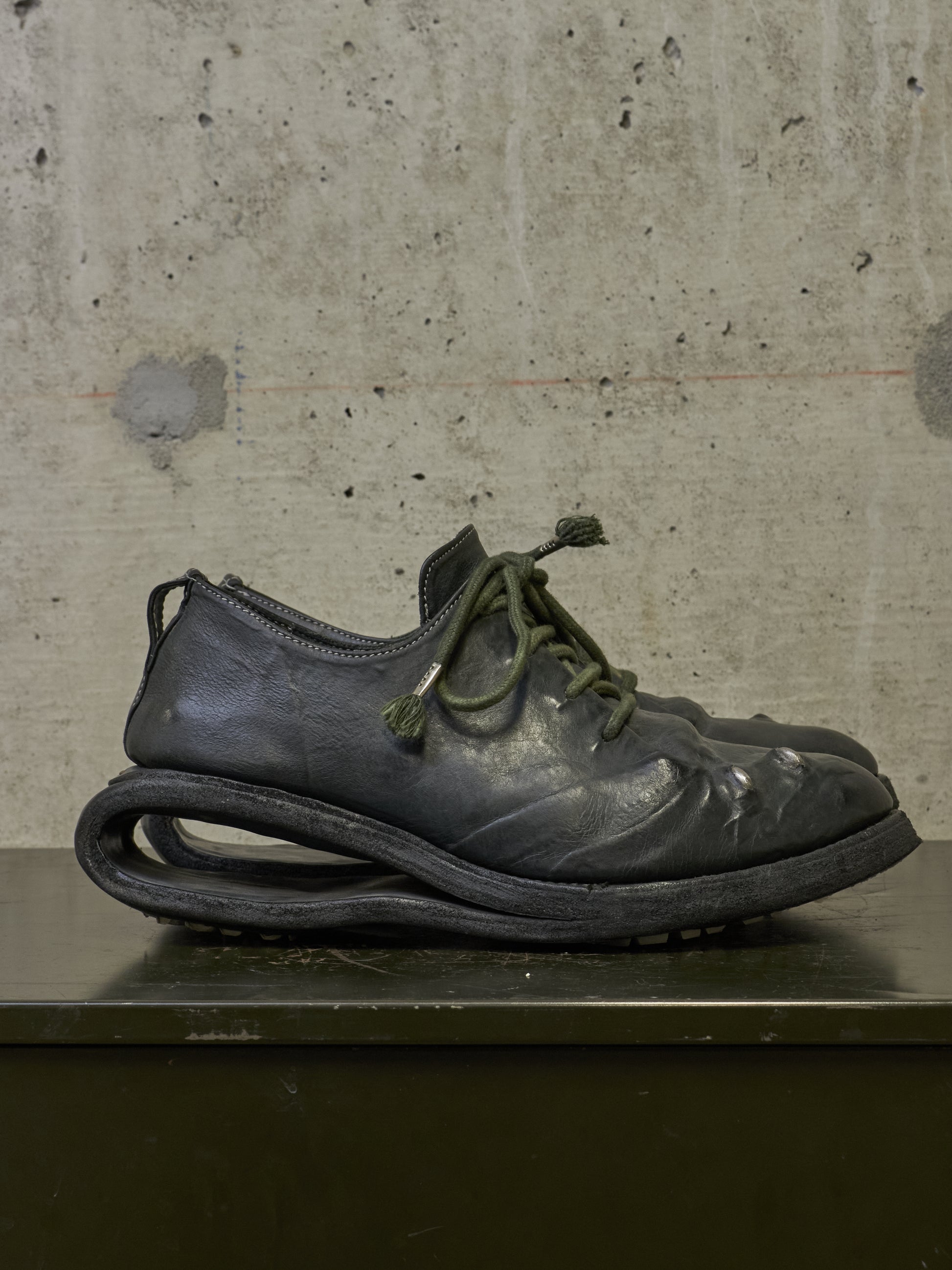 A single dark gray leather shoe with olive green laces and a thick sculptural curled sole, displayed on a dark surface against a textured concrete wall.