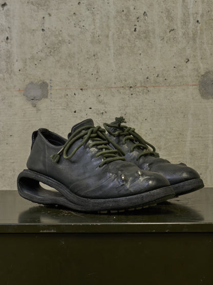 A pair of dark gray leather shoes with unique hollow-cut soles and olive green laces, displayed on a dark surface against a textured concrete wall.