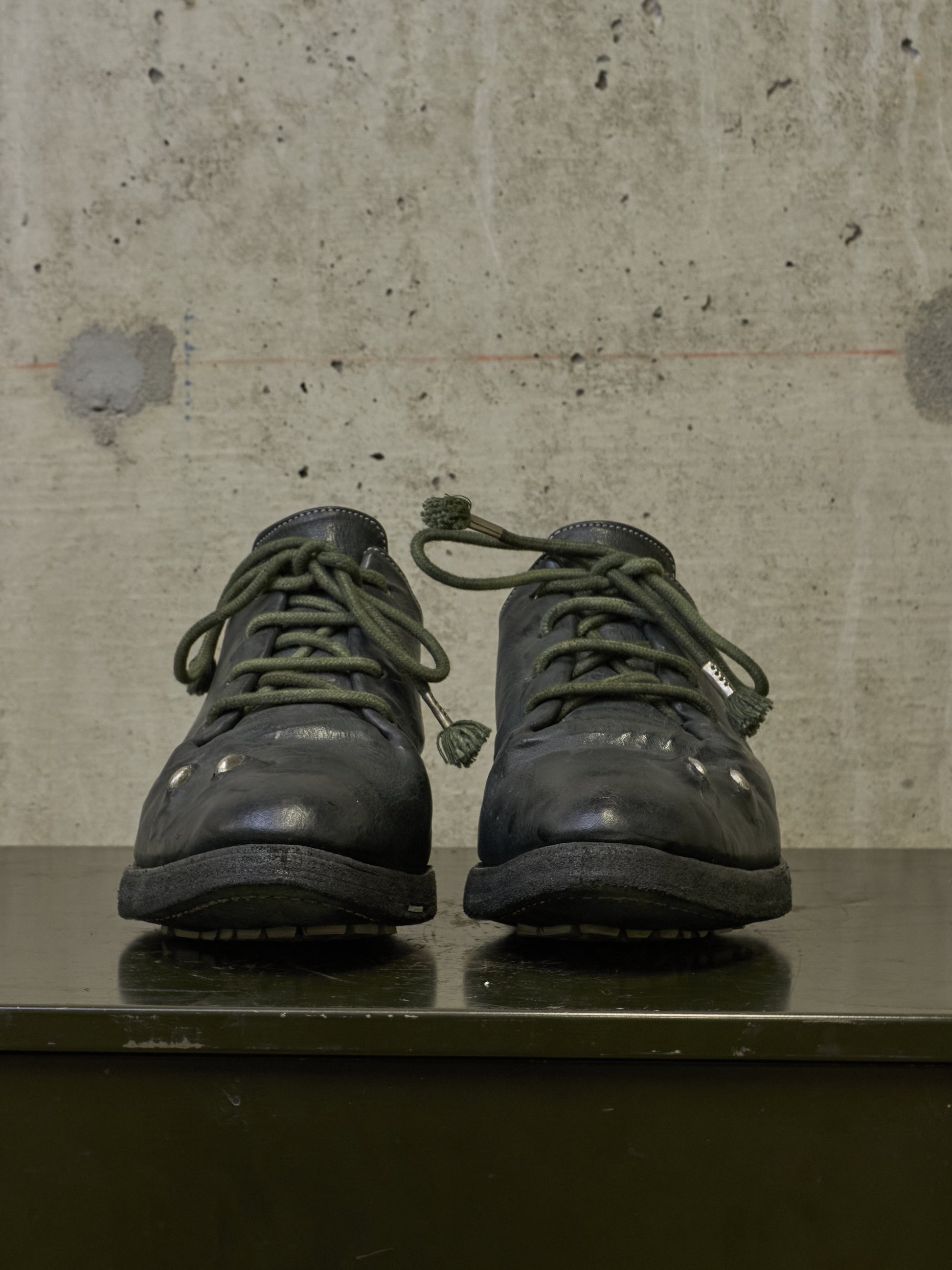 A pair of dark olive green lace-up boots displayed side-by-side on a dark reflective surface against a textured concrete wall.