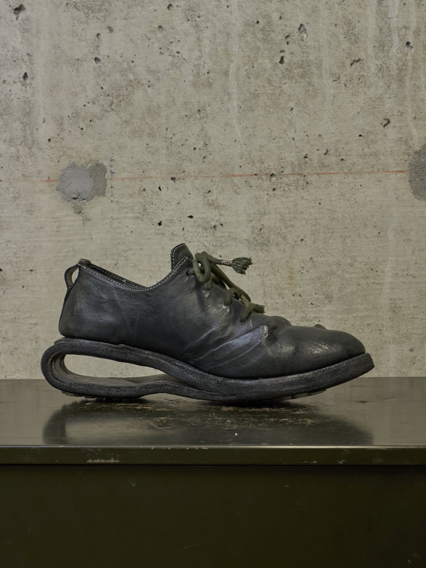 A single black leather lace-up shoe with a distinctive curved sole, resting on a dark surface against a textured concrete wall.