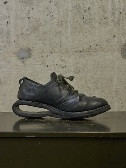 A single black leather lace-up shoe with a distinctive curved sole, resting on a dark surface against a textured concrete wall.