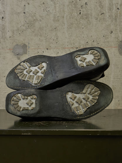A pair of black leather shoes shown from the sole side, resting on a dark surface against a textured concrete wall.