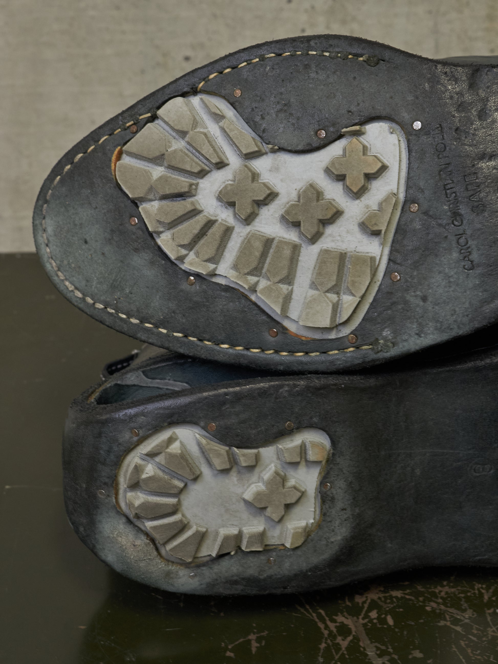 Close-up of the outsoles of a pair of dark gray leather shoes stacked on a dark reflective surface, showing the tread pattern and hand-written markings.