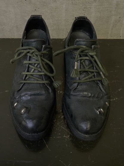 A pair of worn black leather lace-up shoes with olive green laces, photographed from above on a dark surface against a textured gray background.