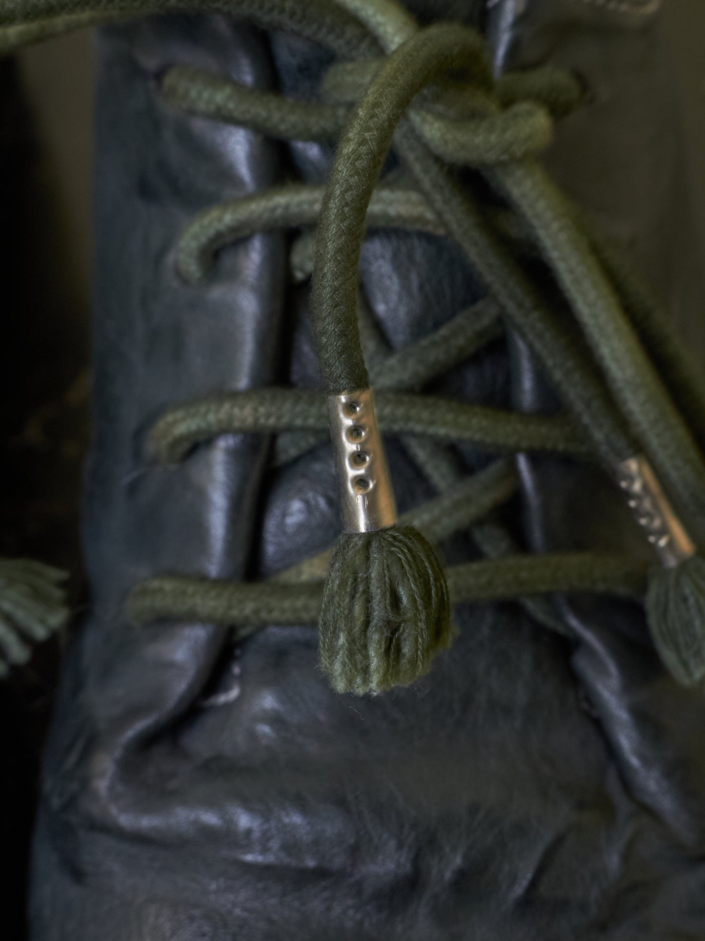 Close-up of olive green rope laces threaded through a black leather lace-up boot, with metal eyelets visible.