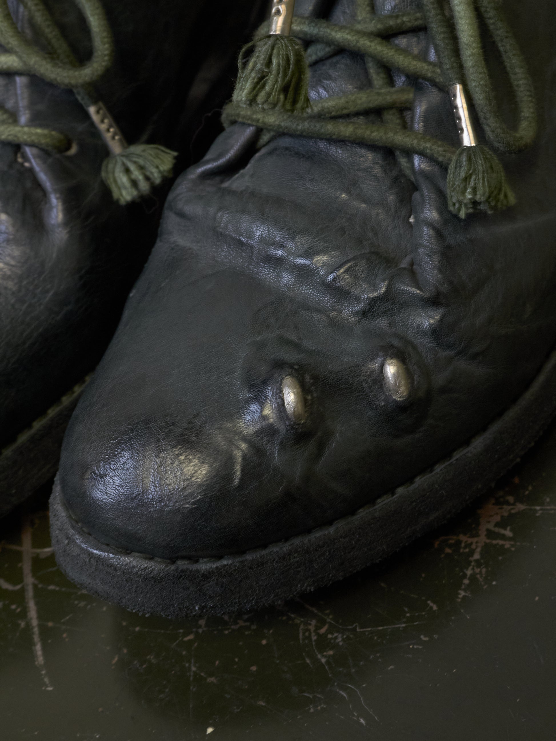 Close-up of a black leather lace-up boot with green laces and decorative tassels, resting on a dark surface.