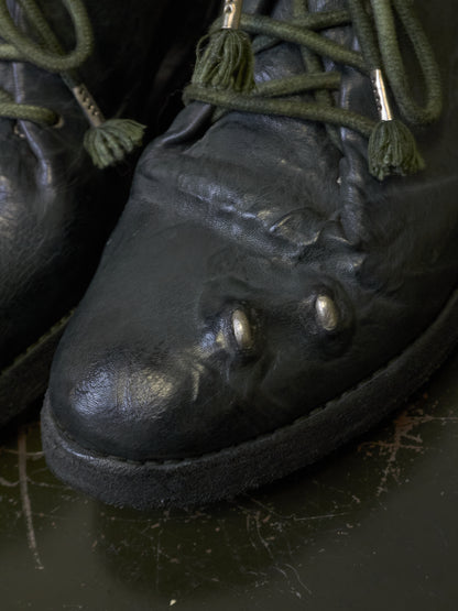 Close-up of a black leather lace-up boot with green laces and decorative tassels, resting on a dark surface.