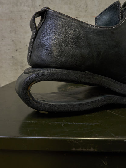 A close-up of a black leather shoe heel and sole, resting on a dark green surface against a textured concrete wall.