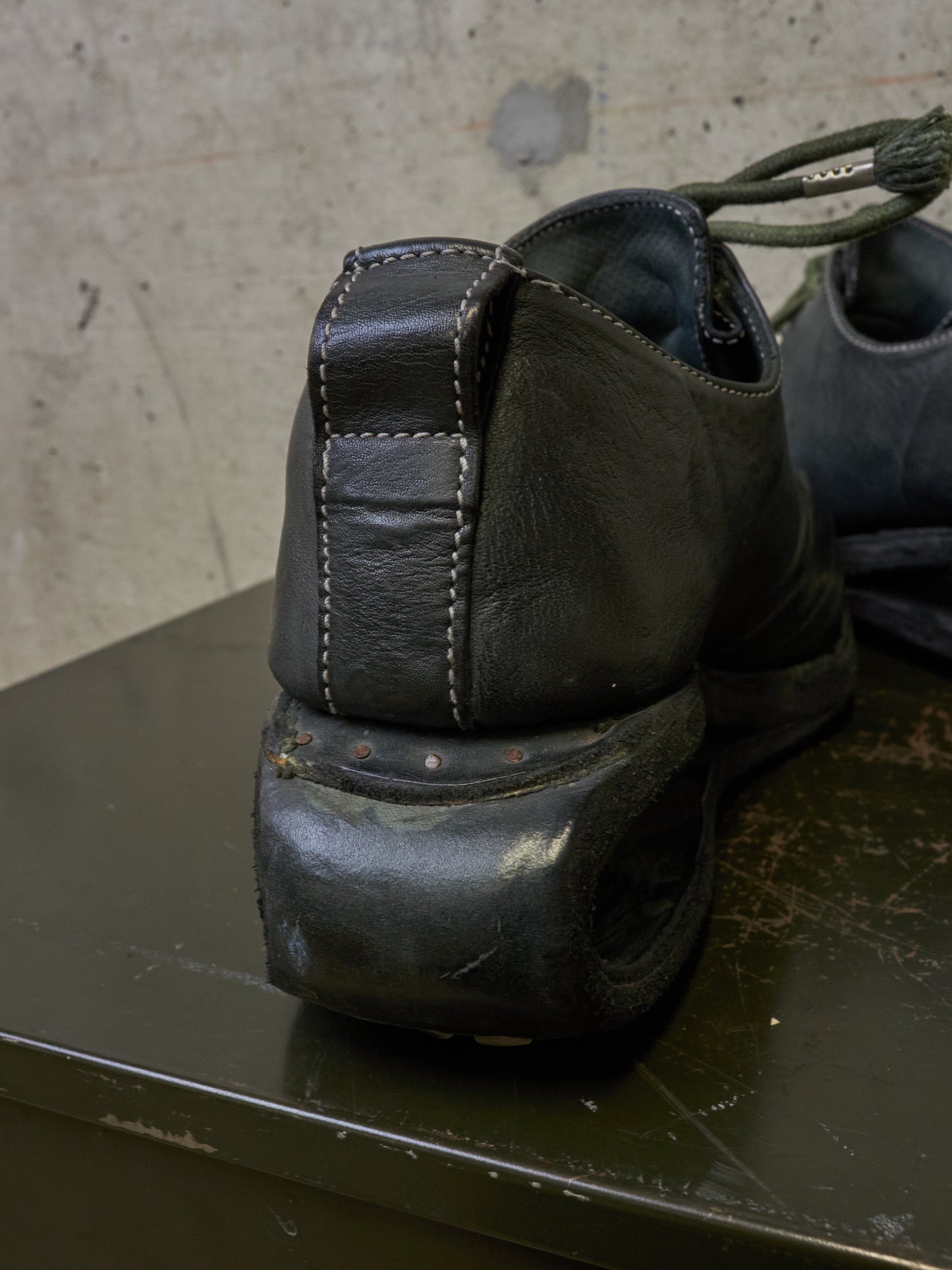 A close-up of a black leather boot or shoe heel, showing the back of the upper and sole, resting on a dark surface against a textured concrete wall.