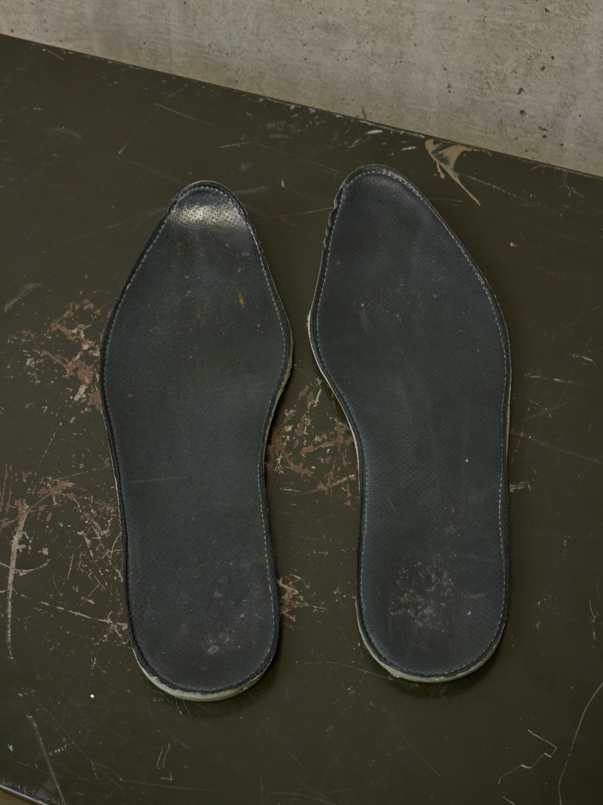 A pair of black leather shoe insoles displayed flat on a dark, scratched surface against a concrete wall.