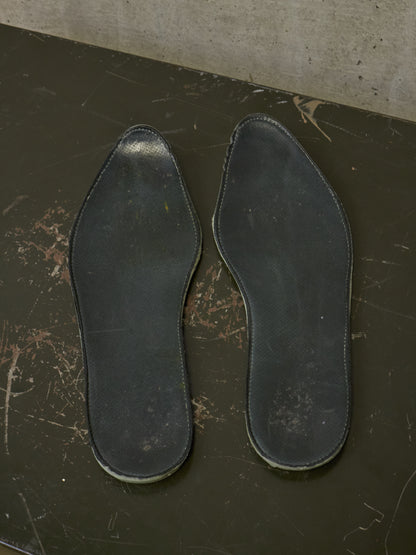 A pair of black leather shoe insoles displayed flat on a dark, scratched surface against a concrete wall.