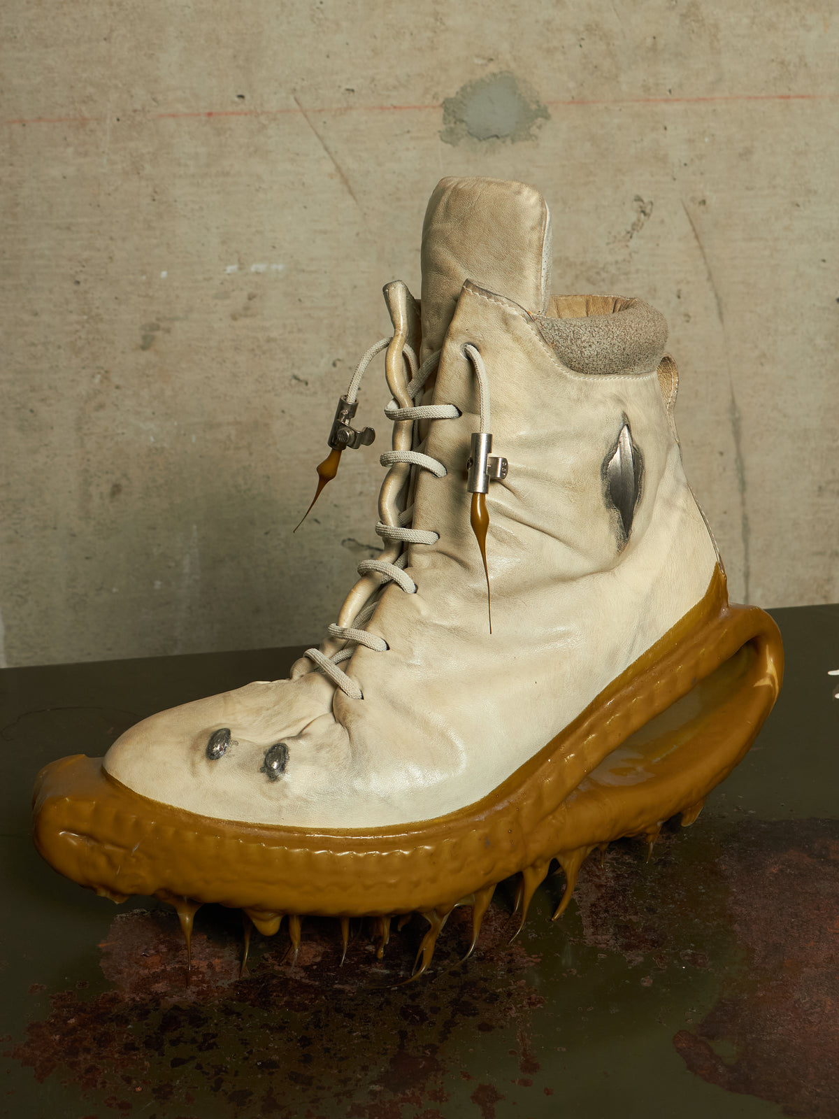 A surreal, horror-themed high-top boot sculpture featuring a beige leather upper with white laces, a metallic silver side accent, and a dripping, organic brown sole resembling a creature's mouth with sharp teeth, set against a textured concrete background.
