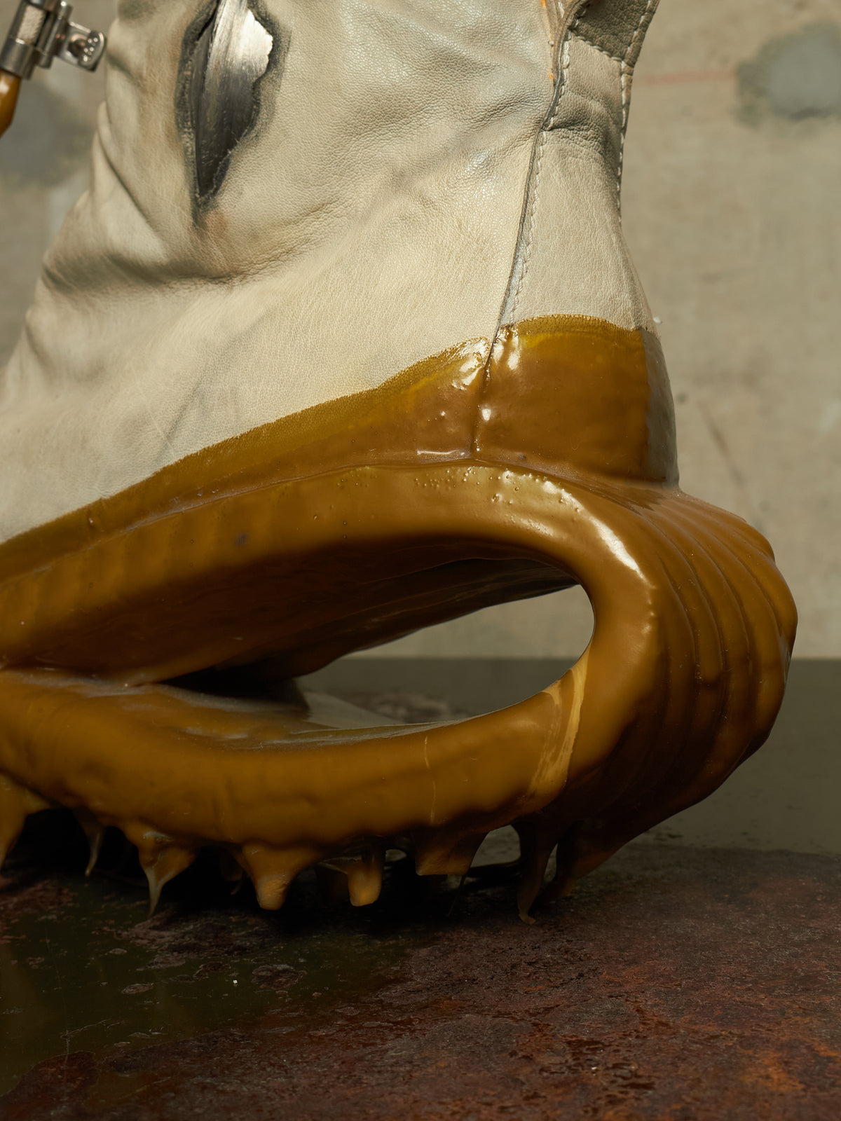 A close-up of a white leather boot with a glossy amber-brown sole or outsole, photographed against a rustic industrial background.