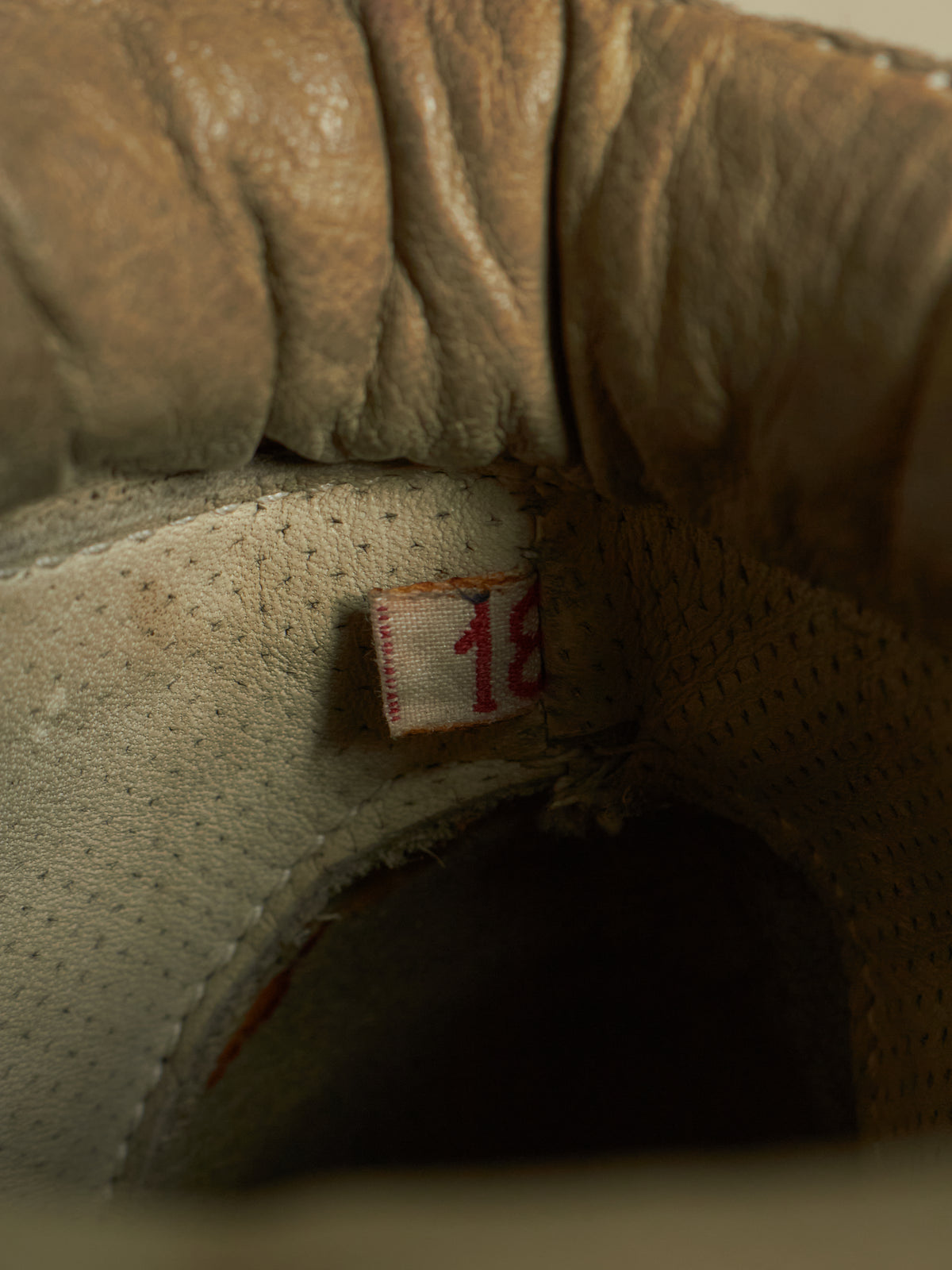 Close-up of the interior opening of a worn tan leather shoe or boot, showing textured leather, stitching, a small fabric tag with red numbering, and a dark shadowed interior.
