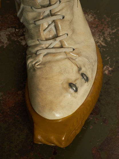 A close-up of a beige leather lace-up boot with a thick, glossy mustard-yellow sole, photographed against a dark, textured, rusty surface.