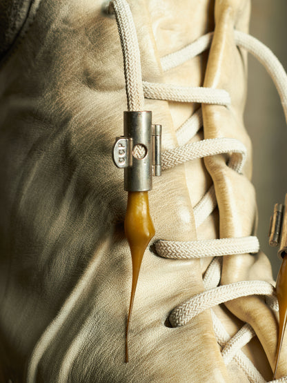 Close-up of a beige leather lace-up shoe with thick white laces and a unique amber-colored silicone drop accent attached near the laces.