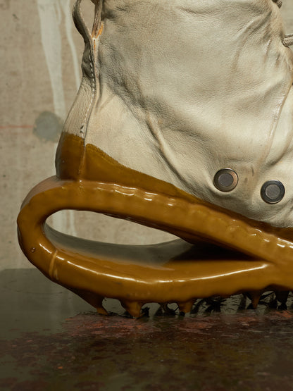 A close-up of a beige leather boot with a glossy amber-brown sculptural sole or outsole, photographed against a textured neutral wall and dark reflective surface.