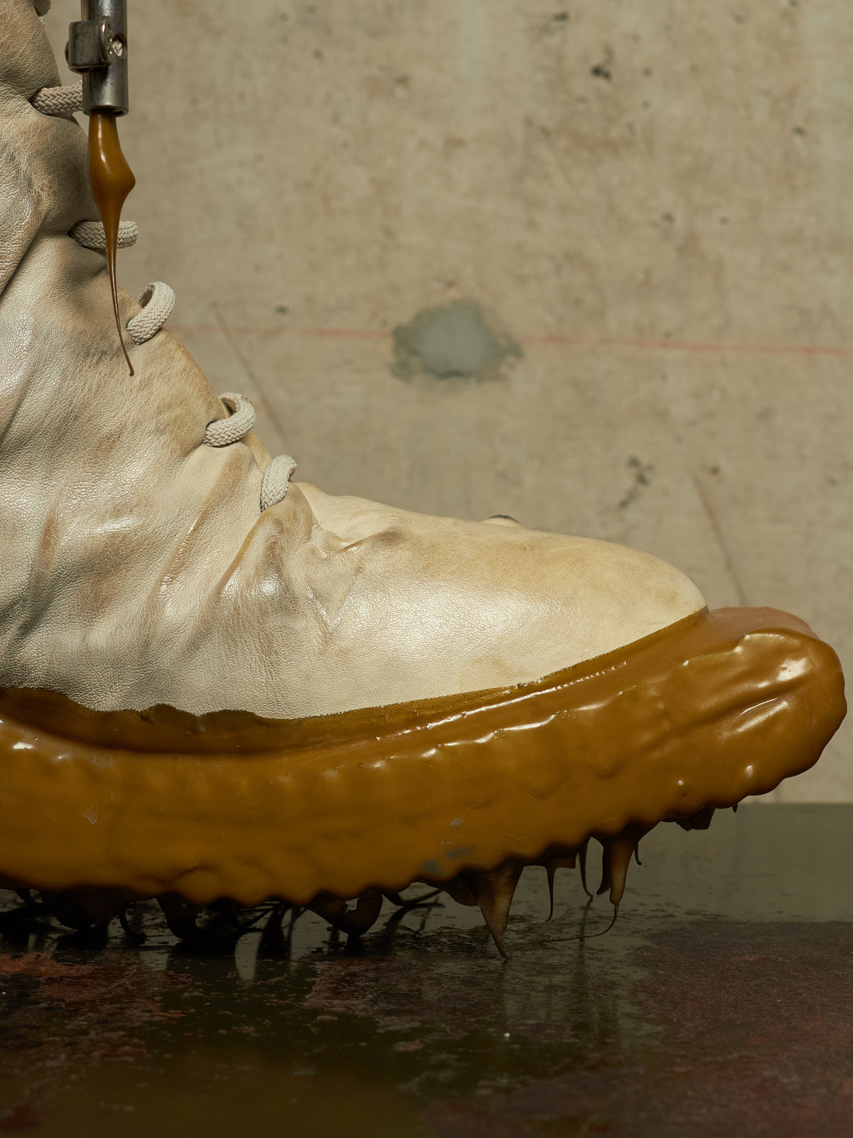 A close-up of a beige leather boot with a thick, glossy brown substance dripping from the sole, set against a textured concrete background.