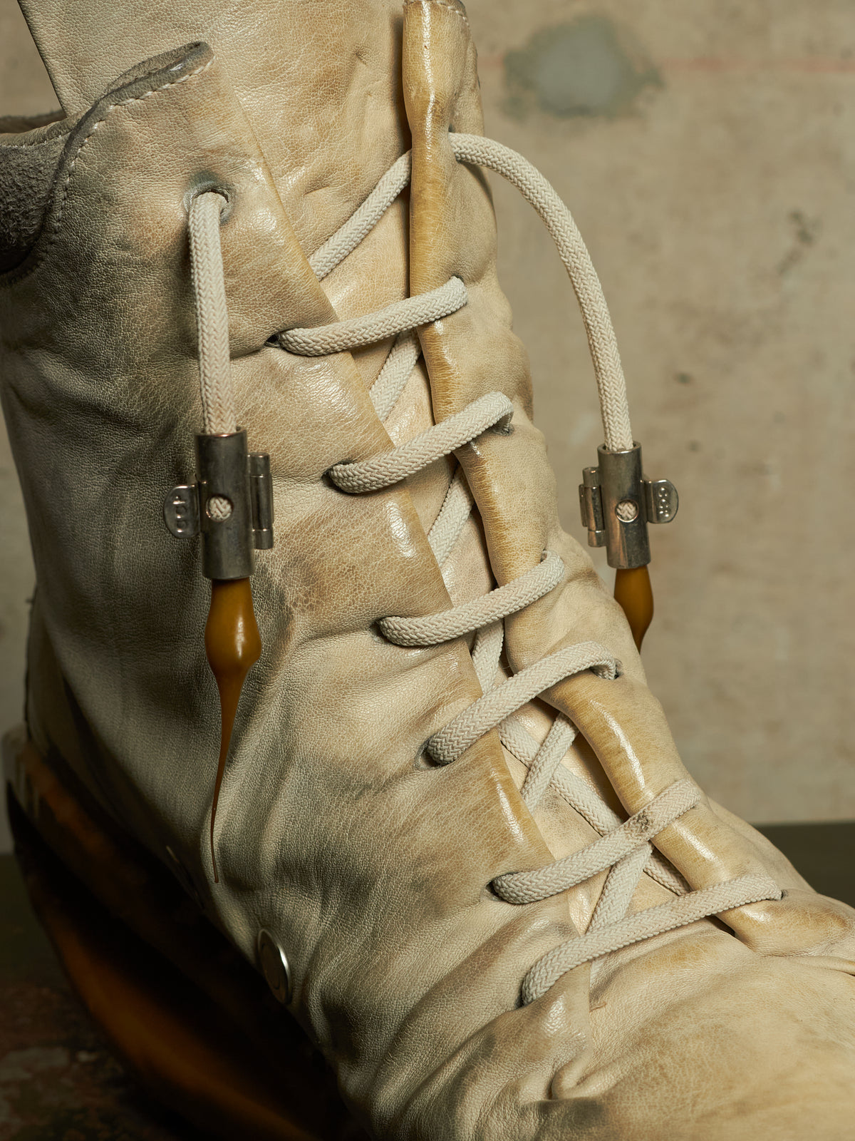 A close-up of a beige leather lace-up boot with thick off-white laces, amber-colored loops, and dark metal slider hardware, set against a textured concrete background.
