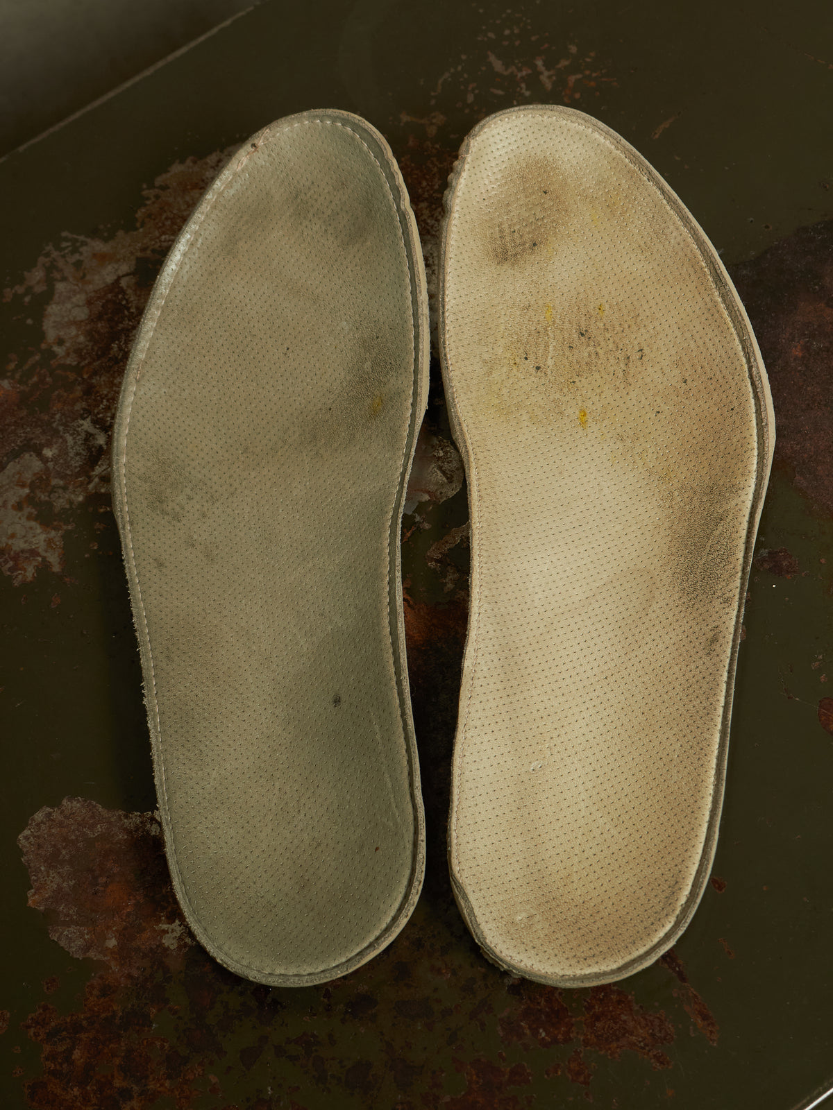 A pair of used, worn-in orthopedic shoe insoles displayed side-by-side on a dark, rusty metal surface.