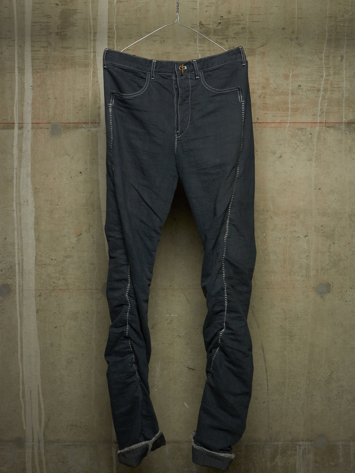 Dark charcoal gray slim-fit jeans with distinctive light gray contrast stitching, hanging on a wire hanger against a textured concrete wall.