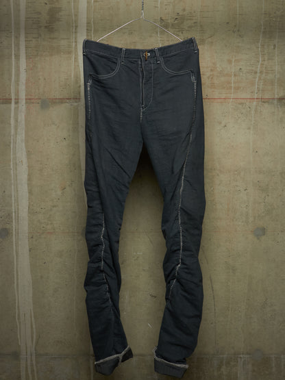 Dark charcoal gray slim-fit jeans with distinctive light gray contrast stitching, hanging on a wire hanger against a textured concrete wall.