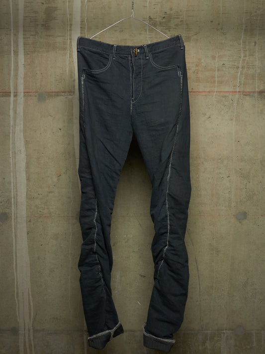 Dark charcoal gray slim-fit jeans with distinctive light gray contrast stitching, hanging on a wire hanger against a textured concrete wall.