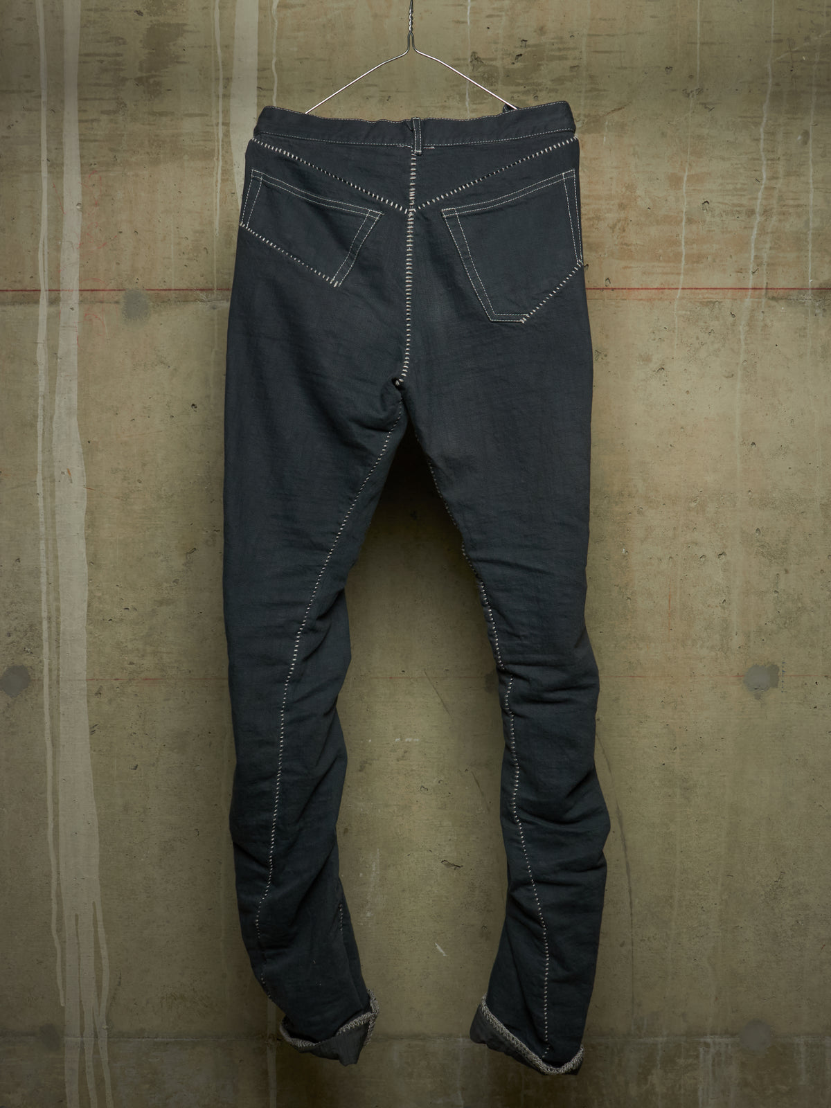 Dark charcoal gray denim pants hanging on a wire hanger against a textured concrete wall, showing the back pockets and contrast white stitching.