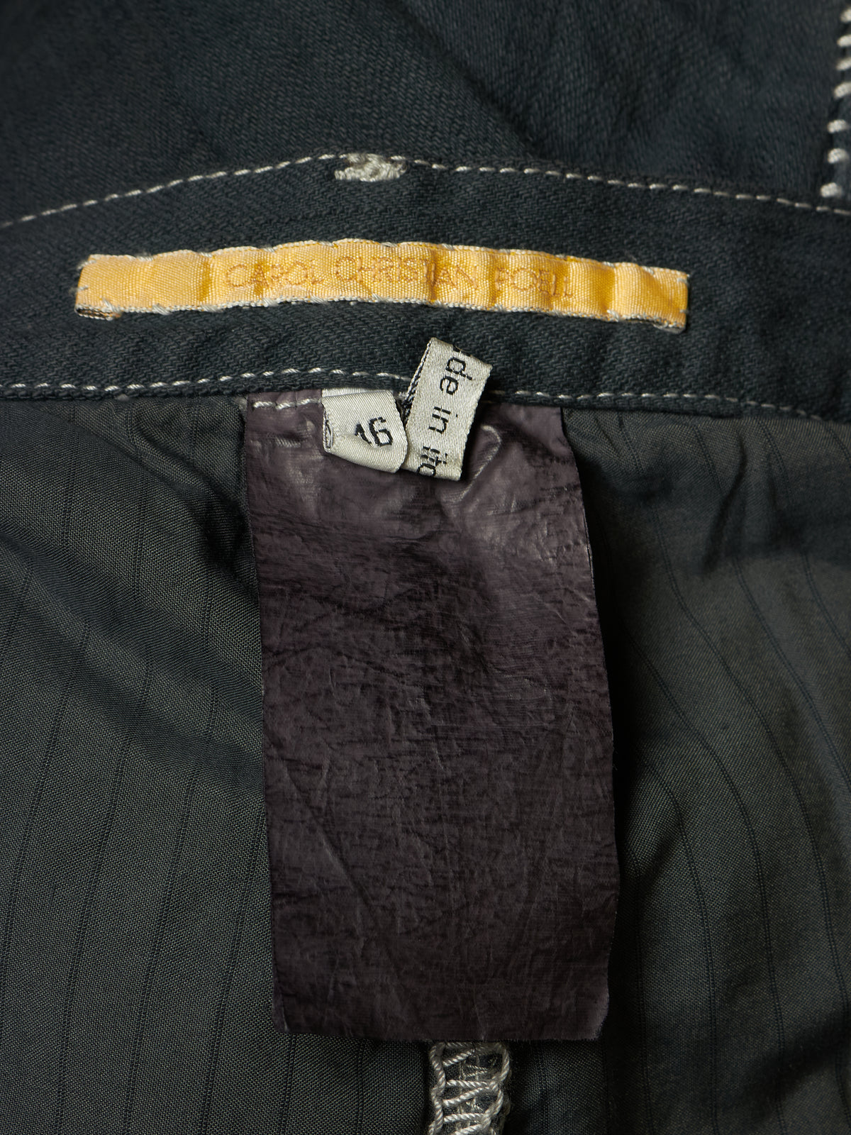 Close-up of the interior label and waistband of a dark charcoal gray pinstriped garment, showing a yellow woven size label and a white 'made in Japan' tag.