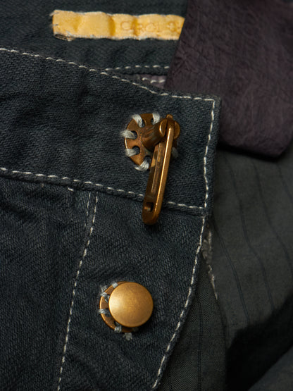 Close-up detail of dark denim fabric with white contrast stitching, a brass snap button, and a decorative metal clasp, alongside a dark purple textile.
