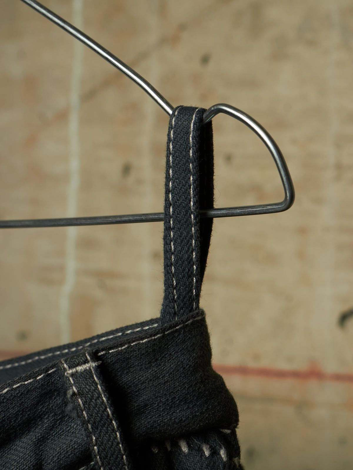 Close-up detail of dark denim fabric with white contrast stitching hanging from a metal hanger against a textured beige background.