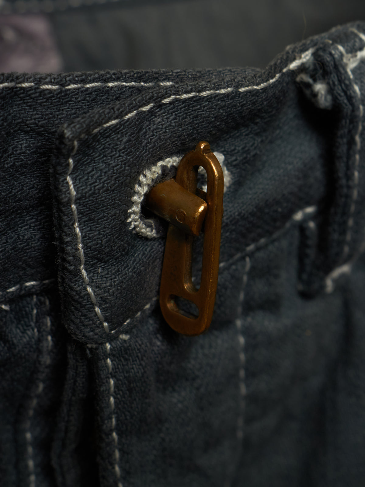 Close-up of dark indigo denim fabric with white contrast stitching and a small bronze metal loop or hardware detail near the waistband.