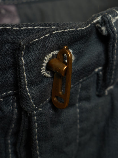 Close-up of dark indigo denim fabric with white contrast stitching and a small bronze metal loop or hardware detail near the waistband.