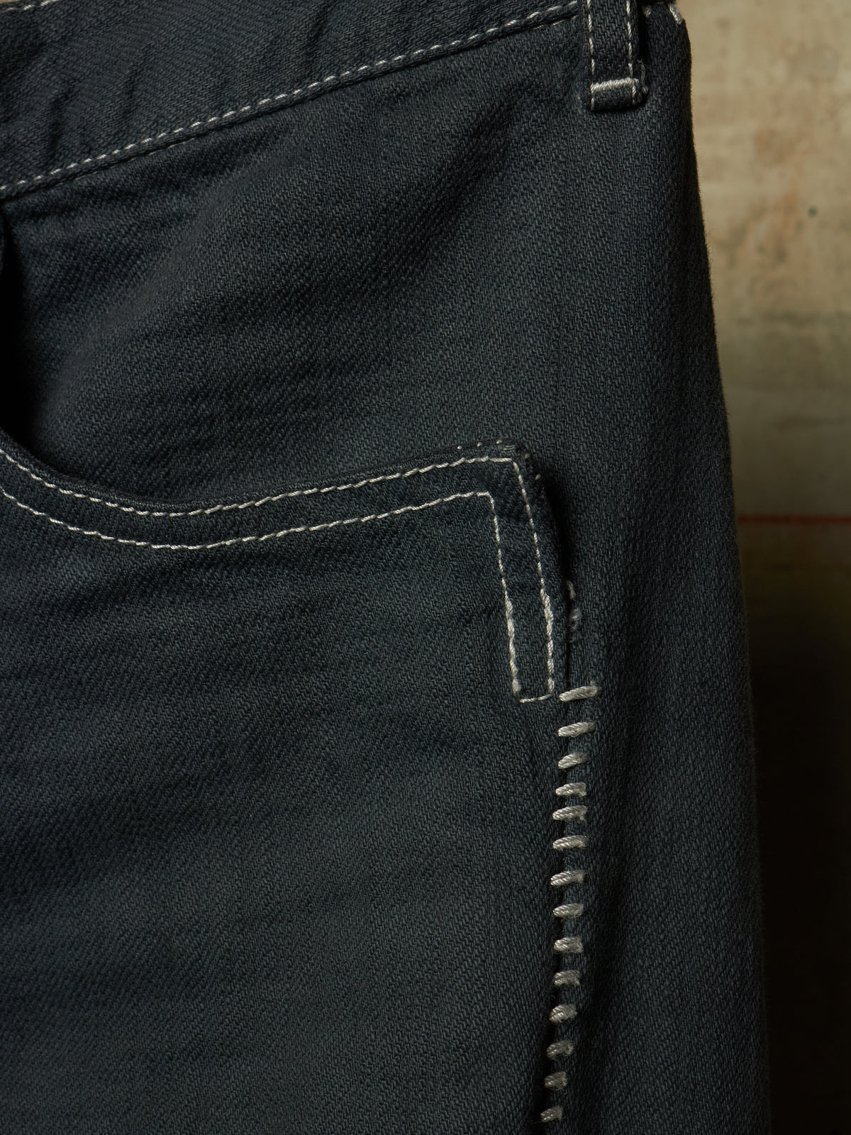 Close-up of the back pocket of dark indigo denim jeans, showing white contrast stitching and the texture of the denim fabric.