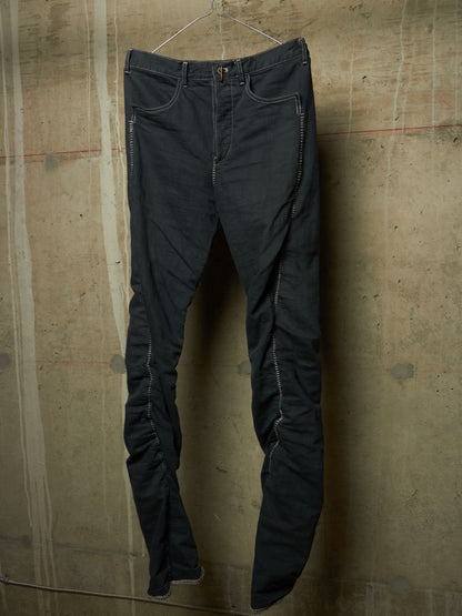 Dark charcoal gray slim-fit jeans with distinctive silver-toned metal rivets along the seams, hanging on a wire hanger against a textured concrete wall.