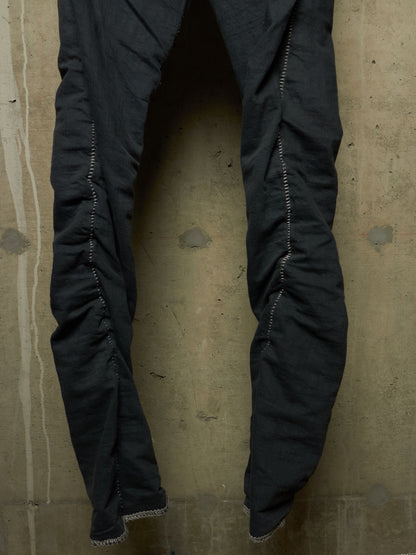 Black crinkled fabric pants with distinctive zigzag stitching down the sides, hanging against a textured concrete wall.
